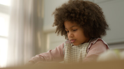 Cute little African American girl in apron adopted child kid ethnic daughter cooking alone in cozy kitchen home make dish bakery family cook prepare dough for cookies buns baking homemade pastries