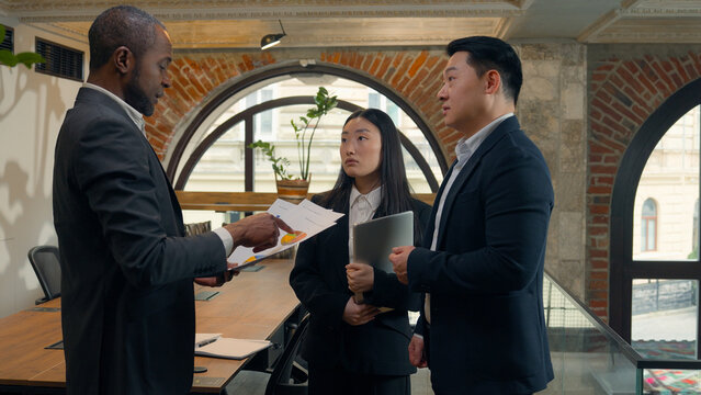 Three multiracial business colleagues coworkers multiethnic in office Asian woman businesswoman Korean Japanese Chinese men businessmen African boss leader talk document discuss pointing data diagram
