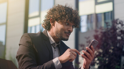 Hispanic happy smiling excited Arabian male Indian amazed businessman receiving good news achievement victory offer opportunity business success achieve man smartphone overjoyed mobile phone in city