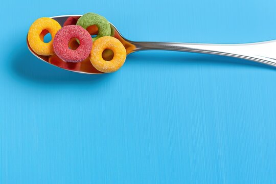 Vibrant scene on blue background with colorful cereal pieces. Silver spoon holds red apple, yellow banana, green apple, and blueberry. Lively and playful composition. - Powered by Adobe