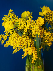 Bouquet of vibrant yellow mimosa and daffodils in a glass vase against a deep blue background, creating a striking contrast and a fresh spring mood