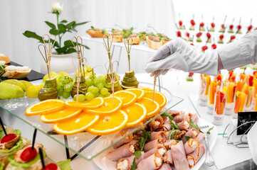 A buffet table with assorted appetizers, including sliced oranges, canapés, and skewered fruits. A hand reaches for an item, showcasing a variety of catering options at an event