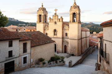Historic stone church with twin bell towers and ornate facade, located in a charming village with stone buildings and red-tiled roofs, surrounded by scenic hills.