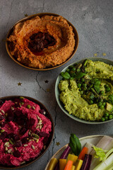 Plates with colorful hummus with pea, beetroot, sun-dried tomato flavors and fresh vegetables on gray background top view close up