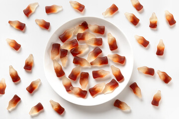 Gummy jellies in shape of soda bottles and full of candy white plate on white background. View from above.