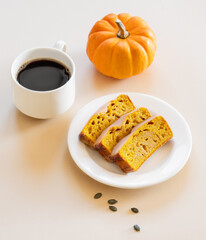 Slices of pumpkin sweet bread or cake with icing on a plate on a light background with decorative pumpkin and a cup of coffee.