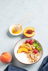 Greek natural yogurt with oat granola or muesli, peach, raspberry and honey in a bowl on a blue background with fresh fruits, berries and napkin.