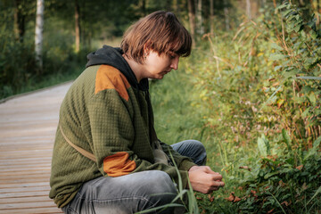 A teenage boy is sitting on the edge of a wooden path surrounded by greenery, wearing a green and orange jacket. He gazes thoughtfully into the distance, immersed in the natural environment.
