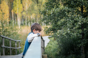 Fototapeta premium A young boy leans on the railing of a wooden bridge and looks thoughtfully into the distance. Surrounded by greenery and trees, the scene captures a quiet, thoughtful moment in a natural setting.