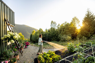 A woman waters her garden in the early morning, surrounded by lush greenery and a stunning mountain...