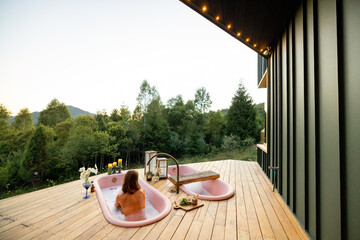 A serene moment of relaxation as a woman enjoys a bubble bath in a pink bathtub on a wooden deck, surrounded by the tranquil beauty of nature at sunset