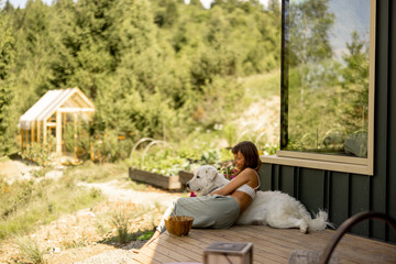A serene scene where a woman and her loyal dog relax on the porch, taking in the beauty of the natural landscape. The reflection in the glass adds depth to the composition
