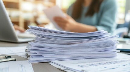 A cluttered desk with a stack of papers and a person reviewing documents.