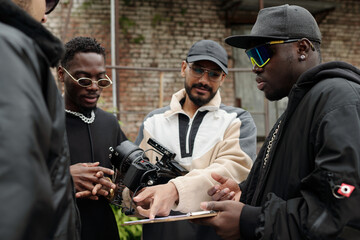 Group of young creatives examining film equipment and discussing project outdoors. Focused members...