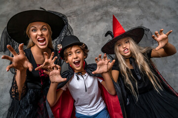 Obraz premium Happy family having fun scaring with look in camera standing over grey background. Mother and children kids in witch vampire costumes and makeup on a celebration of Halloween on dark grey background