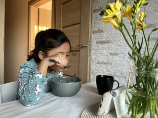 Asian little girl watching mobile phone while eating dinner in the modern kitchen. Screen time health risks concept