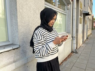 A smiling Asian woman in a hijab uses her smartphone near building in Europe. Beautiful Muslim woman connects with friends and family through her smartphone.