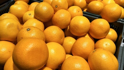 A close-up photo of a pile of bright orange in a black plastic crate. Fresh and juicy oranges, with visible dimples and a slightly textured rind.