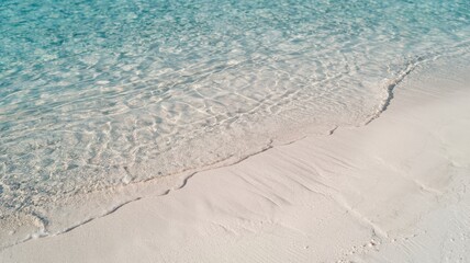 A photo of a pristine beach with clear turquoise waters. The water's edge is characterized by a gentle wave lapping onto the sandy shore. The sand appears soft and fine, with subtle patterns and ridge