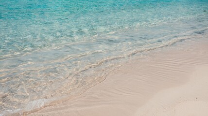 A serene photo of a pristine beach with clear turquoise waters. The water's edge is characterized by a gentle wave lapping onto the sandy shore. The sand appears soft and fine, with subtle patterns