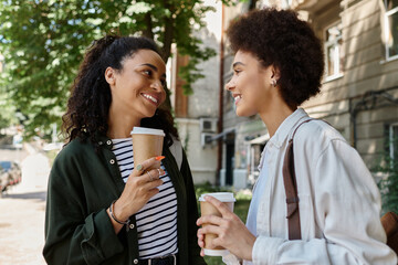 Two joyful women share laughter and caffeine while exploring the outdoors in their vibrant neighborhood.