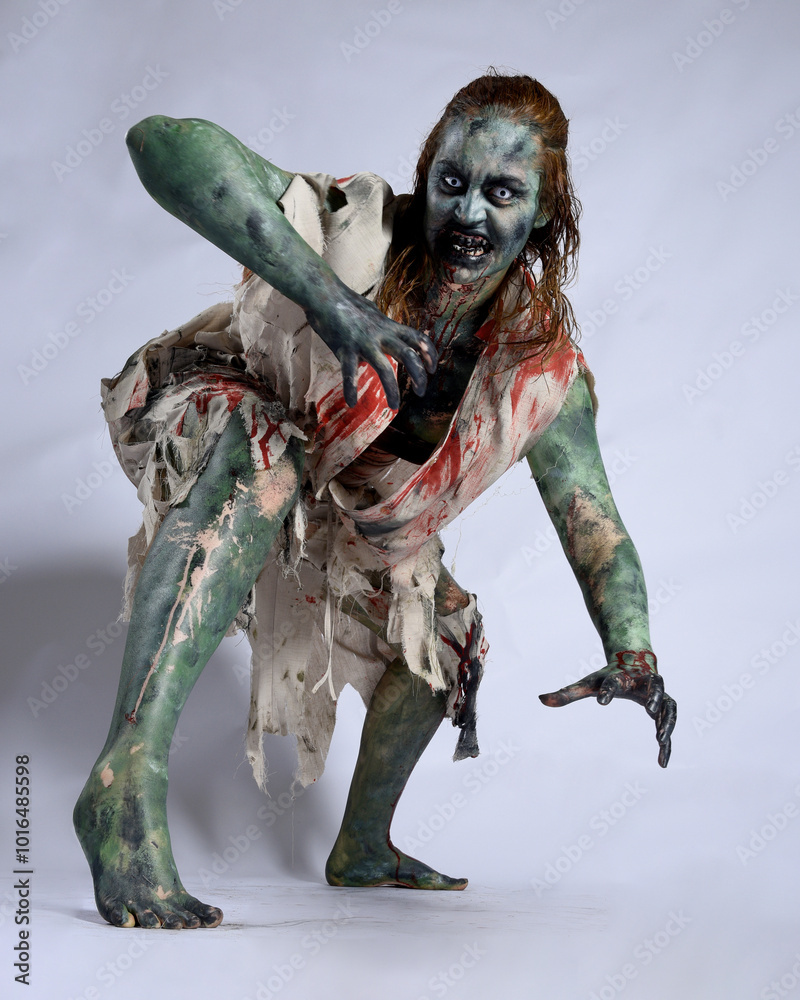 Wall mural portrait of scary female model wearing spooky halloween costume, ripped clothes and fake blood like an undead demon apocalyptic zombie character. Isolated figure, crouching pose dark studio background