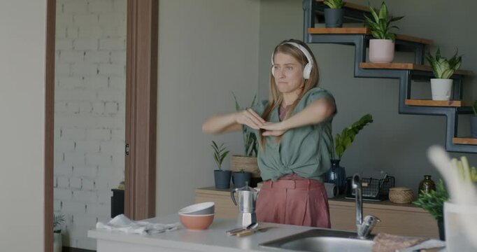 Happy girl wearing headphones listening to music and dancing moving to pop song having fun in kitchen at home. Millennial culture and youth concept.