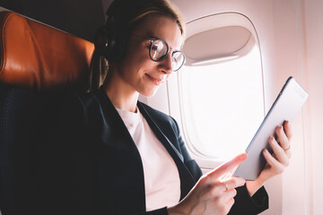 Positive young woman sitting in passenger seat in airplane and reading interesting book via...