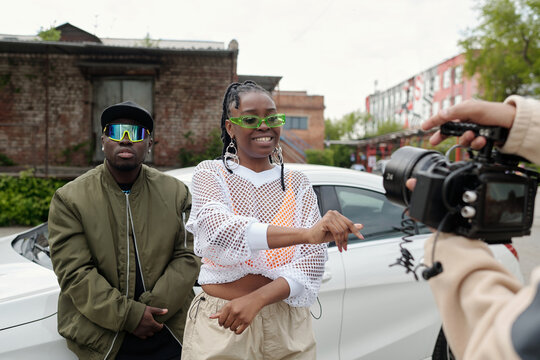 Two people standing outdoors behind parked car, one interviewing other using camera with microphone, both wearing sunglasses, casual attire, urban backdrop visible