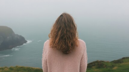 Fototapeta premium Küstenruhe: lockige Brünette im Pullover mit Blick auf den nebligen Ozean für saisonale Reflexion