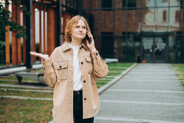 Fototapeta premium A surprised blonde woman talking on the phone outdoors. Urban lifestyle concept. Beautiful woman walking on the city street. People emotion