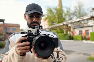 Obraz premium Man wearing hat and glasses holding professional camera while standing outdoors, adjusting settings with focused expression, blurry background of buildings and trees adding depth