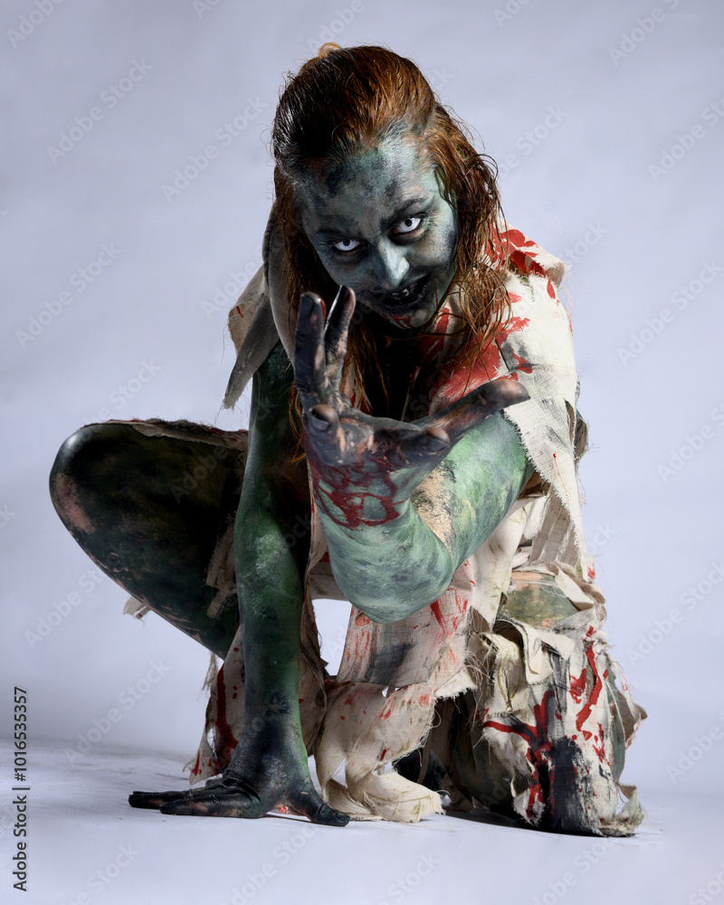 Poster portrait of scary female model wearing spooky halloween costume, ripped clothes and fake blood like an undead demon apocalyptic zombie character. Isolated figure, crouching pose dark studio background