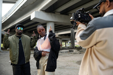 Two people posing under urban overpass with third person filming them using professional camera equipment focused on capturing dynamic expressions in gritty city environment