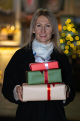 Young smiling blonde woman in a black coat with gift boxes looks at the camera on the street in the evening. Concept of holidays, Christmas. Vertical photo