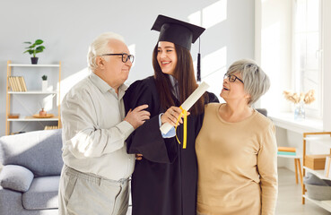 Happy and proud elderly parents are congratulating adult graduate daughter in mortarboard and gown,...