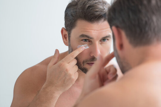 Man applying cosmetic face cream on mirror. Facial treatment. Portrait of man with bare naked shoulders touching skin. Sexy man with skin care product. Male face creme. Perfect skin, morning routine.