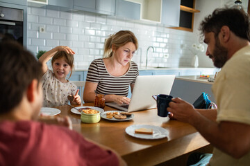 Mother working on laptop while family has breakfast in kitchen