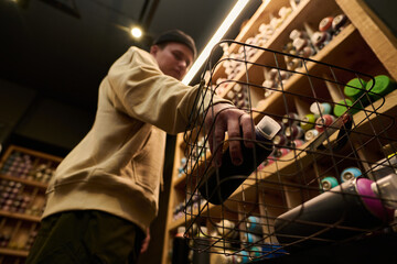 View from below of hand of young man putting cans with graffiti paints into basket or shopping cart while buying goods