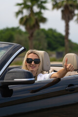 Portrait of a young smiling blonde woman in sunglasses driving a convertible convertible showing a thumbs up. Concept of travel, vacation, rental. Vertical photo