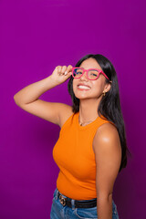  Cheerful woman wearing bright pink glasses and orange top, posing against vibrant purple backdrop