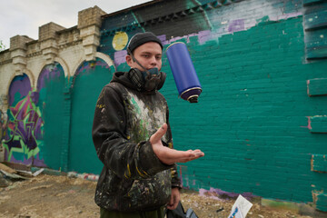 Guy in respirator, beanie cap and hoodie throwing blue can with paint while standing against green...