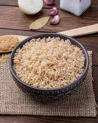 Cooked brown whole rice in a bowl over wooden table with onion, bay leaf and garlic