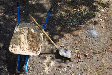 A shovel and a wheelbarrow with sand in the shade of a tree, construction work on the ground, the...