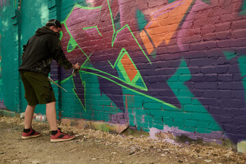 Young man in casualwear bending forwards and spraying purple paint on brick wall of building while working over graffiti artwork © pressmaster