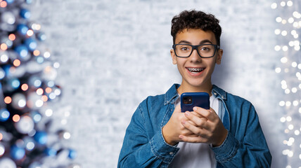 Excited surprised shocked astonished happy funny young man wear braces glasses spectacles open mouth hold typing cell phone cellular smartphone cellphone. Christmas xmas x-mas tree lights background.
