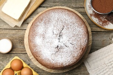 Tasty chocolate sponge cake with powdered sugar and ingredients on wooden table, flat lay