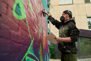 Guy in casual attire and protective mask standing in front of wall of building and painting graffiti artwork in the city