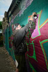 Side view of young man skilled in graffiti painting spraying green paint from can on brick wall while creating new artwork