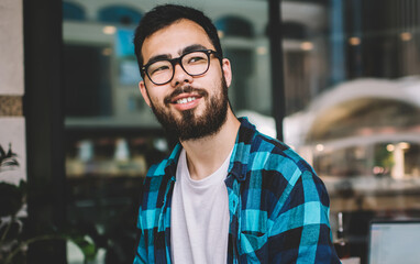 Happy male blogger in optical eyewear for vision correction pondering on idea for university essay, asian hipster student holding textbook for education during time for autodidact in street cafeteria